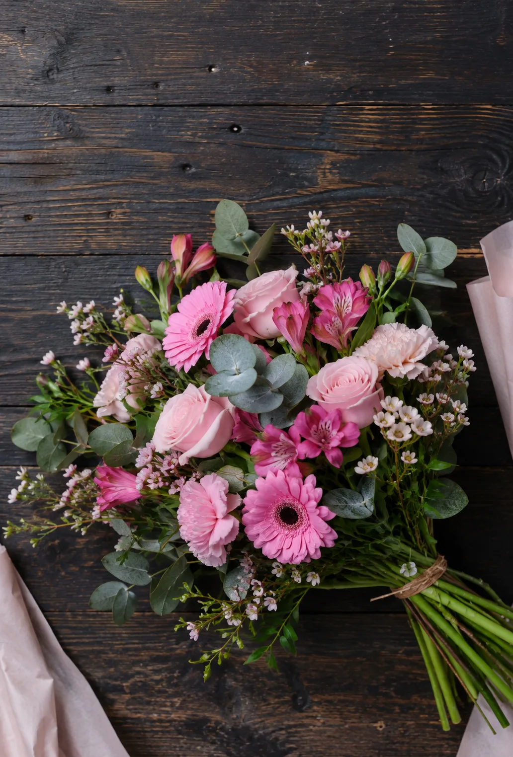 A wrapped bouquet of pink roses, gerberas and eucalyptus on a dark wood bench